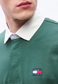 Κοντινό πλάνο ενός ατόμου που φοράει πράσινο πόλο μπλουζάκι με λευκό γιακά και ορατό λογότυπο Tommy Jeans στο στήθος.