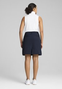 Γυναίκα που στέκεται με την πλάτη στραμμένη, φοράει λευκό αμάνικο τοπ, σκουρόχρωμη φούστα μέχρι το γόνατο και λευκά αθλητικά παπούτσια σε γκρι φόντο.