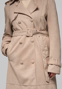 Beżowy dwurzędowy płaszcz trencz z miękkiej tkaniny, z paskiem w talii, dużymi guzikami i bocznymi kieszeniami.