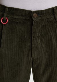 Pantalons en velours côtelé vert olive avec des côtes verticales, comportant un bouton marron et un détail en boucle métallique rouge sur la ceinture.