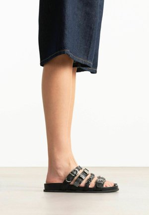 Bas des jambes portant un pantalon court en jean foncé et des sandales plates noires avec plusieurs brides bouclées.