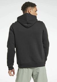 Hombre con cabello corto y rizado, vistiendo una sudadera negra y pantalones cortos de color verde claro, visto desde atrás contra un fondo liso y claro.