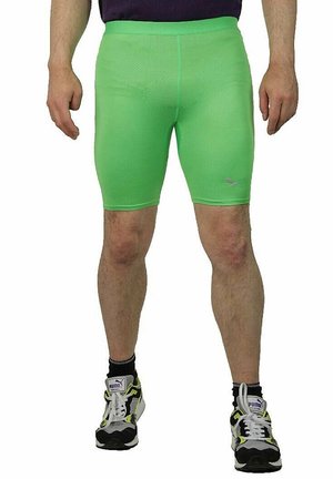 Mann trägt leuchtend grüne enge Sportshorts und schwarze Puma-Sneaker mit weißen Schnürsenkeln, steht mit entspannten Armen seitlich am Körper.