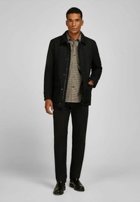 Man in een zwarte jas, geruite bruine shirt, zwarte broek en zwarte schoenen, staande met één hand in zijn zak tegen een grijze achtergrond.