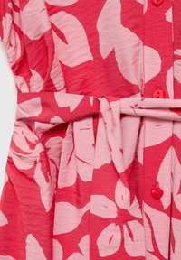 Stoff mit rot-pinken Blumenmuster, Knöpfen und einem gebundenen Gürtel Detail an einem Kleidungsstück.