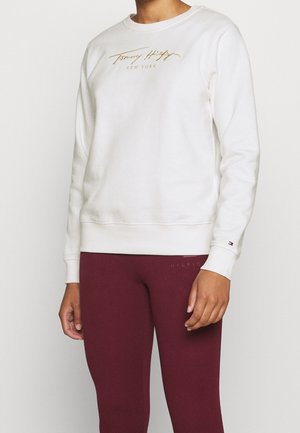 Osoba ubrana w białą bluzę Tommy Hilfiger z długim rękawem z logo w kolorze złotym oraz bordowe legginsy.