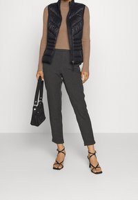 Gestepptes schwarzes Westchen, braunes geripptes Langarm-Oberteil, hoch taillierte graue Hose, schwarze Handtasche, schwarze Riemchen-Sandaletten mit Absatz.