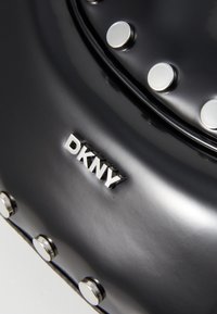 Sac à main noir brillant avec ferrures argentées et clous ronds. Présente un logo DKNY proéminent sur le devant. Design élégant et moderne.