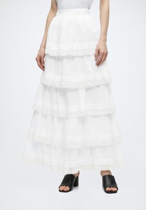 Frau trägt einen gestuften weißen Maxirock mit Spitzenbesatz und schwarze Blockabsatz-Sandalen mit offenen Zehen, steht vor einem schlichten hellen Hintergrund.