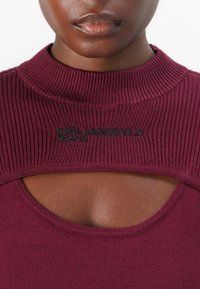 Bordo, pletena majica z visokim ovratnikom in izrezom na prsih. Črn logo "KARL LAGERFELD JEANS" jasno izpostavljen.