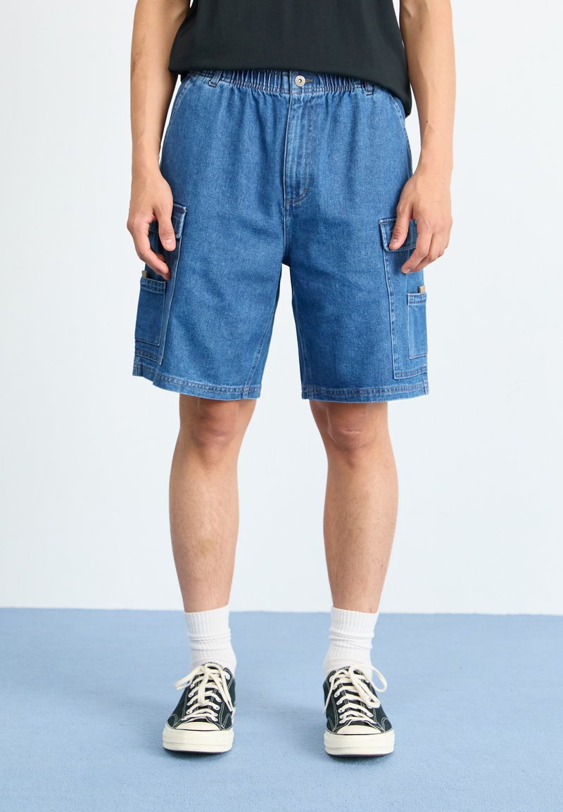 Shorts din denim cu talie elastică, cu buzunare laterale, de culoare albastru deschis și o croială lejeră. Purtați cu adidași negri și ciorapi albi.