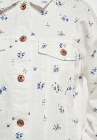 Hvit denimjakke med blå blomsterprint, kobberknapper og to brystlommer. Distinktiv søm gir tekstur og detaljer.