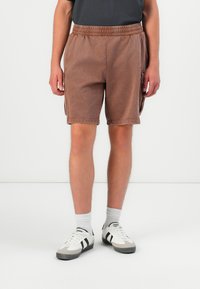 Person iført brune cargo-shorts, hvide sokker og hvide sneakers med sorte striber, stående foran en ensfarvet lys baggrund.