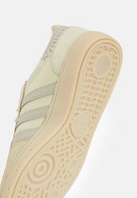Beiger Sneaker mit strukturiertem Wildlederobermaterial, drei kontrastierenden Streifen und einer Gummisohle mit geometrischem Profil.