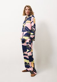 Kleurrijke lounge-outfit met een marineblauwe achtergrond en grote roze, gele en groene bladpatronen; losse blouse met bijpassende broek.