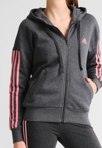 Sweat à capuche gris à fermeture éclair, fabriqué en tissu doux, avec une capuche, des cordons de serrage noirs et des accents roses à trois bandes sur les manches.