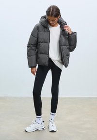 Sivi puffer jakna s kapuljačom, kutijastog kroja, kvadratnog dizajna, nosi se preko bijele majice, uparenih s crnim tajicama i bijelim sportskim cipelama.