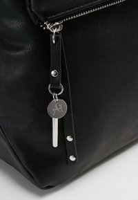 Sac à main en cuir noir avec une finition texturée, doté d'une fermeture éclair, d'accents métalliques et d'un porte-bonheur de marque suspendu à une sangle.
