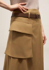 Jupe marron en tissu lisse, arborant des poches superposées et une ceinture marron. Le design se caractérise par des lignes épurées et des détails structurés.