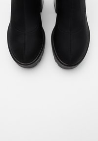 Botas negras hasta el tobillo hechas de un material sintético liso con puntera redondeada, que presentan costuras mínimas y una suela gruesa y texturizada.