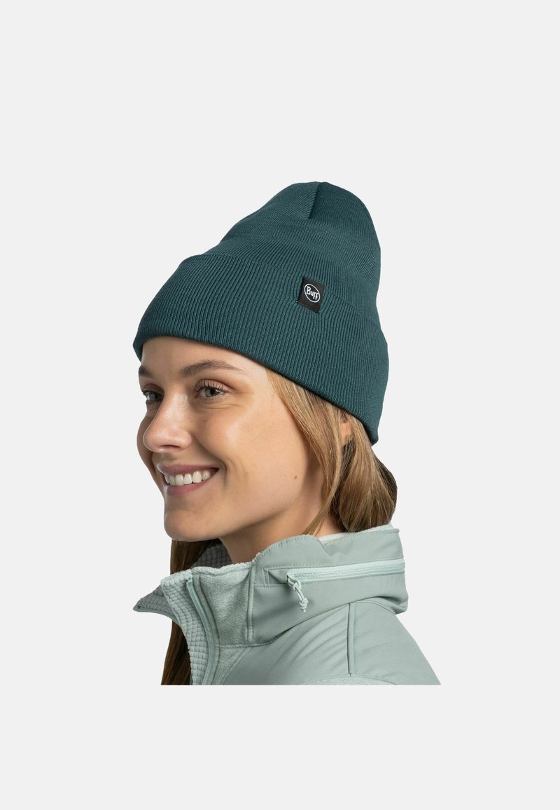 Gorro de punto acanalado en color verde azulado con un parche de logo, que presenta una parte superior puntiaguda y un ajuste ceñido. Se lleva junto a una chaqueta verde claro con cremallera.