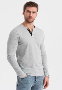 Harmaa pitkähihainen paita Henley-kauluksella, jossa on mustia yksityiskohtia ja kaksi ruskeaa nappia. Päällä siniset farkut.