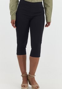 Pantalones negros cortos con una cinturilla elástica, costuras laterales y cremalleras en los tobillos, combinados con sandalias de cuña de esparto en color beige.