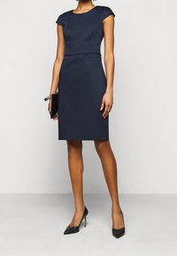 Robe ajustée bleu marine avec des manches courtes, longueur genou, texture lisse et un accent discret à la taille. Associée à des talons aiguille noirs.