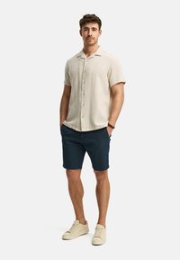 Lys beige kortærmet skjorte med knapper og struktureret stof, parret med mørkeblå shorts og beige sneakers. Enkel, afslappet design.
