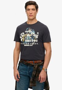 Mørkegrå bomulds T-shirt med et vintage grafisk design i farverig print. Sammen med blå denimjeans og en grønternet skjorte bundet om taljen.