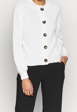 Kobieta w białym prążkowanym kardiganie z dużymi ciemnymi guzikami i czarnych spodniach, jedna ręka w kieszeni, na tle jednolitego jasnego tła.