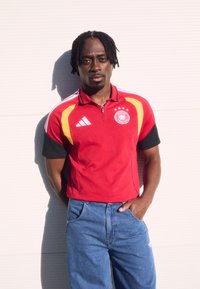 Κόκκινο κοντομάνικο πόλο με μαύρες και κίτρινες λεπτομέρειες, με γιακά και έμβλημα τριών αστεριών, συνδυασμένο με μπλε τζιν παντελόνι.