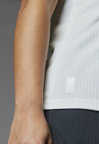 White ribbed tank top with a textured finish, featuring a small white logo patch at the hem. The model is wearing dark, ribbed pants.