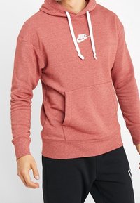 Man i en röd Nike-hoodie med vita dragsnören och svarta byxor, stående med armarna något böjda.