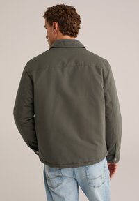 Uomo con capelli ricci che indossa una giacca verde oliva e jeans azzurri, mostrato di spalle su uno sfondo neutro.