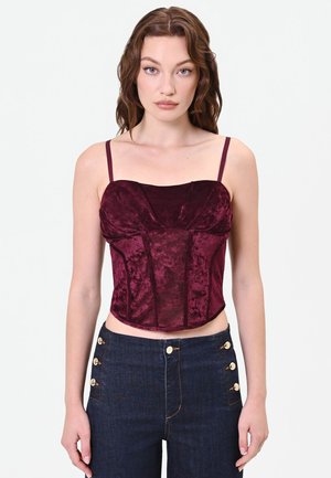 Giovane donna con capelli mossi castani indossa un top corsetto senza maniche in velluto bordeaux e jeans scuri a vita alta con bottoni dorati.