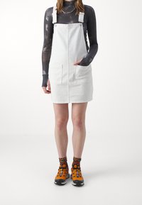 Biała sukienka ogrodniczka z denimu z dwoma przednimi kieszeniami, z regulowanymi ramiączkami. Połączona z pomarańczowymi butami sportowymi i wzorzystymi skarpetkami.