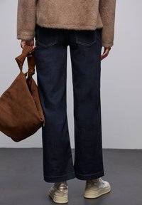 Szerokie ciemnoniebieskie dżinsy z kontrastowym przeszyciem, połączone z beżową, puszystą kurtką. W lewej ręce trzymana jest brązowa zamszowa torba.