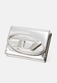 Stříbrná metalická peněženka s výrazným oválným logem. Hladký povrch, skládací design a viditelné detaily šití. Kompaktní tvar.