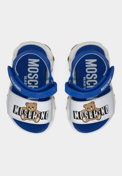 Μπλε και λευκά πέδιλα μωρού Moschino με γραφικό αρκουδάκι και ρυθμιζόμενα λουράκια σε απλό φόντο.