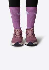 Zapatillas deportivas rosas con parte superior de tela texturizada y cordones morados. Combinadas con calcetines morados hasta la rodilla que presentan un logo blanco y textura acanalada.