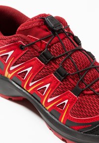 Red athletic shoe with a mesh upper, black laces, and bold white and yellow accents. Features a textured black rubber sole for traction.