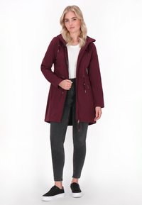 Bordeaux lange jas met een capuchon, touwtje in de taille, ritssluiting en zijzakken. Gecombineerd met donkere jeans en zwarte sneakers.