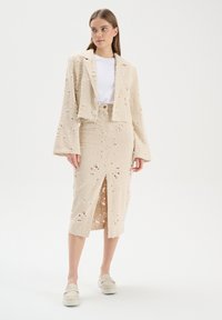 Beige cropped blazer met uitlopende mouwen, gecombineerd met een knie-lange rok met uitgesneden patronen en een voorrits. Witte t-shirt eronder.