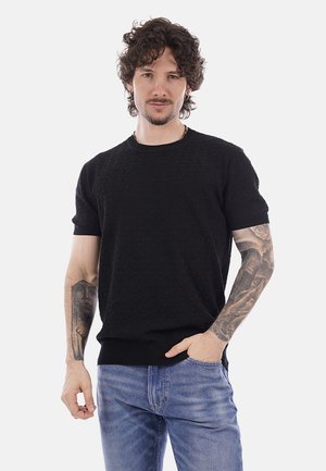 Uomo con capelli ricci che indossa una camicia nera a maniche corte con trama e jeans blu, mostrando tatuaggi dettagliati su entrambi gli avambracci.