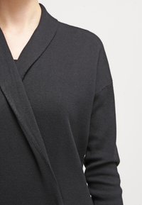 Top lungo a maniche nere in maglia avvolgente con pannelli frontali incrociati indossato da una persona su uno sfondo chiaro e semplice.