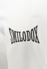 Vit tyg med en slät struktur har svart tryckt text "SMILODOX" i en fet, stiliserad font. Inga ytterligare mönster eller detaljer synliga.
