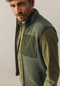 Green fleece vest with a front pocket, zippers, and a soft texture. Worn over a textured olive shirt with button details.