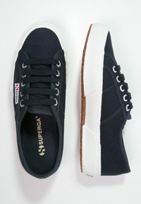 Zapatillas de lona navy con suela de goma blanca y patrón de agarre detallado. Presenta cinco ojales con cordones gris claro y una etiqueta de logo en el lateral.