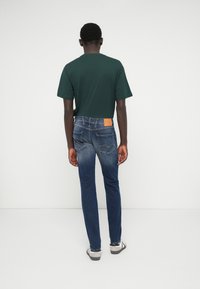 Σκούρο πράσινο βαμβακερό t-shirt με κοντά μανίκια, συνδυασμένο με μπλε τζιν. Το τζιν περιλαμβάνει μια ανοιχτό καφέ δερμάτινη επένδυση στη πίσω μέση και λεπτομέρειες ραφής.